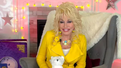 Dolly Parton wears bright yellow jacket and holds plush dog in the set of CBeebies Bedtime Stories