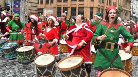 Mick Hall Rows of women in red santa suits and green elf suits sing and play traditional drums with their drumsticks. Old buildings rise up behind.