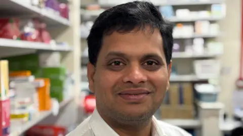 Sridhar Maturi has short black hair and a pale shirt. He is smiling. In the background there are pharmacy shelves.