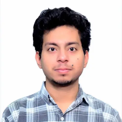 A young man wearing a blue checkered shirt looks into the camera. 
