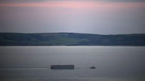 Finnbarr Webster/Getty Images The Bibby Stockholm is seen from a distance, being pulled through the sea by a tugboat. In the background are country hills and a pink sunset.