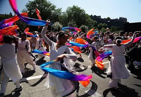 Performers in flowing white outfits dance through a street parade while twirling long ribbons in bright shades of blue, purple and orange. Spectators line the route, with trees and historic buildings visible in the background.
