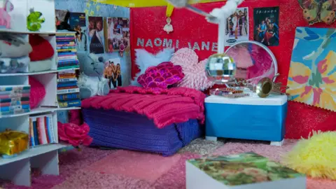 A bedroom with bright pink, red, yellow and blue walls, carpet and bed, with cushions, cuddly toys, posters and books