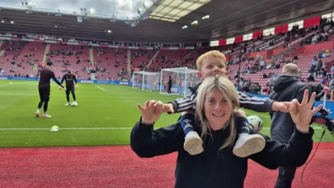 Katie stands at the edge of a football pitch, with Ruairi on her shoulders. Both are smiling. 