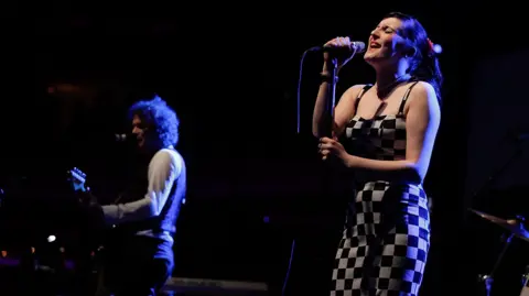 Getty Images Codeine Velvet Club perform onstage - Lou Hickey is singing while wearing a black and white patterned dress, while Jon Lawler plays guitar in the background