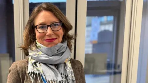 A woman with glasses, in a brown jacket, white shirt and blue scarf stands in a reception room smiling at the camera. 