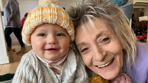 Baby grinning with woolly hat on and an older woman grinning next to him 