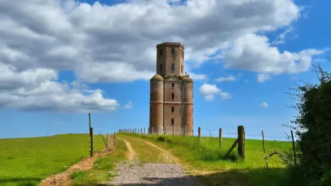 Shaun A large six storey brick tower stands at the end of a gravel track at the top of a grassy hill. The sky above is blue with white clouds and it is a sunny day. The track is lined with a post and wire fence.