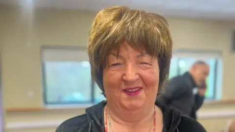 Image of Frances Hamilton smiling at the camera. She is wearing a black zip-up hoodie and a red beaded necklace. She also has pink lip-stick. In the background a man can be seen talking on his mobile phone.