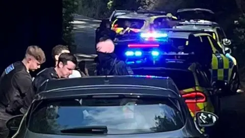 Two police officers in black uniforms standing to the left of a black car. Behind the black car are two police cars, with blue-and-yellow markings and blue lights shining.
