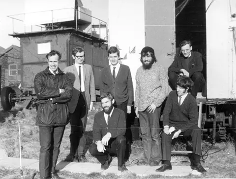 A group of men pose for a photograph in front of buildings in a grassy field. 