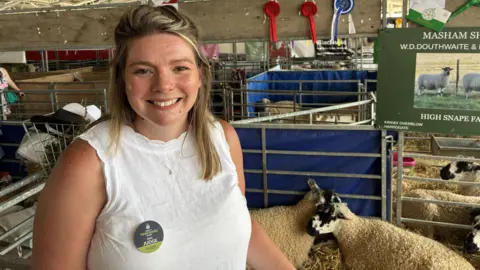 A woman with shoulder length blonde hair stands in front of several sheep pens. She wears a white sleeveless top and a round badge that reads 'Judge'.