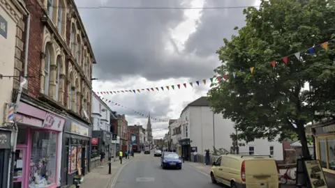 Google Fisherton Street in Salisbury, Wiltshire. Shops and pubs can be seen either side of the street. Cars can be seen travelling along the road whilst a van is parked up on the kerb. Bunting is hanging, stretching across the road. 