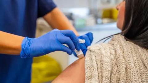 A woman with long brown hair, wearing a beige sweater with the sleeved rolled up. She is sat down and a medical professional wearing blue scrubs and blue latex gloves putting a plaster on the woman's arm. 