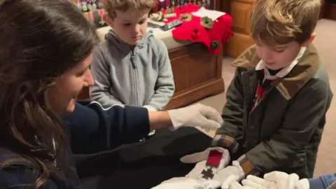 A woman with long brown hair passes a Victoria Cross medal to two young children. They are all wearing white gloves to protect the medal.