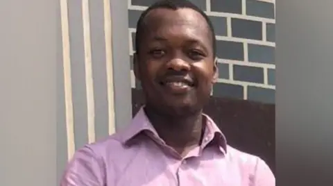 Bedfordshire Police Patrice Che - a man with short black hair wearing a pink shirt and smiling. He is standing in front of a grey brick wall.