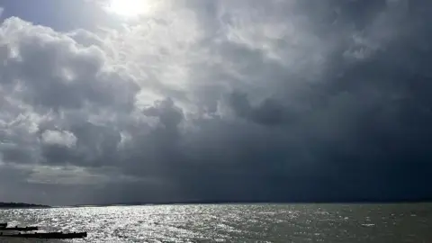 A bright, white sun shines through a break in dark grey clouds above a glistening sea in this dramatic seascape at Hill Head Beach