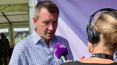 Stuart Anderson MP, a man with short greying hair and wearing a blue and white checked shirt, being interviewed by BBC Radio Shropshire inside a gazebo with a purple backdrop