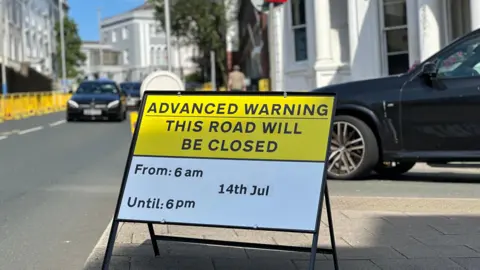 A sign reading 'ADVANCED WARNING THIS ROAD WILL BE CLOSED' From 6am until 6pm - 14th July. In the background are blurred cars driving along a road lined by yellow cones