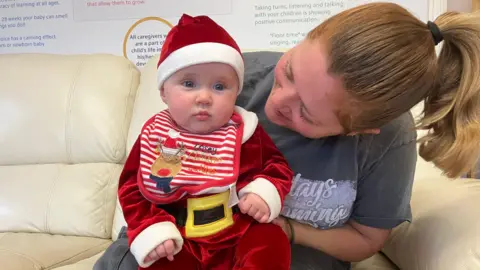 A baby wearing a red and white santa hat, a santa suit with a faux black and yellow, chunky belt, and a red and white stripy bib, which includes an image of a reindeer with a red nose. To the right is a woman with brown hair, who is turned towards the baby. She is wearing a dark t shirt with light-coloured writing on the front.