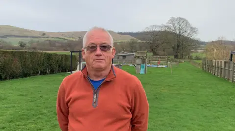 Parish Councillor Ian Tuckett stands in his garden unsmiling on a grey afternoon, with a view to Roundway Down in the background. He is wearing glasses and an orange zipper pullover.
