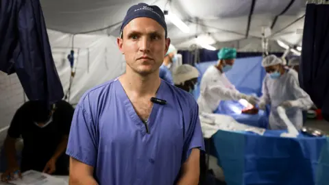 Dr Matt Newport faces the camera but looks to his right, wearing his blue scrubs and a blue surgical cap, with a number of medics who have been blurred in the background of a field hospital in Gaza. 