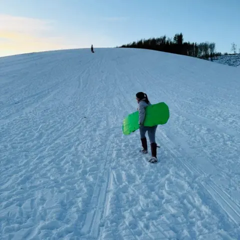 Robyn Fowlie Robyn Fowlie walking up a snow-covered slope with a bright green sledge under her arm, there are trees at the top of the hill.