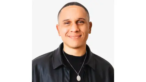 Thomas Morgan A man wearing a black leather jacket and a silver necklace smiles at the camera