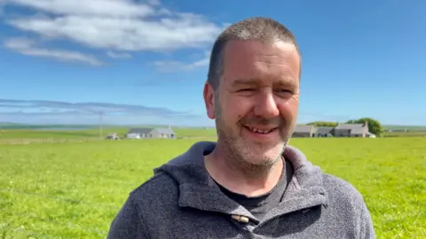 A man with short cropped grey hair and stubble in a grey hooded-top squints into the sunshine, against a backdrop of blue sky and green fields.