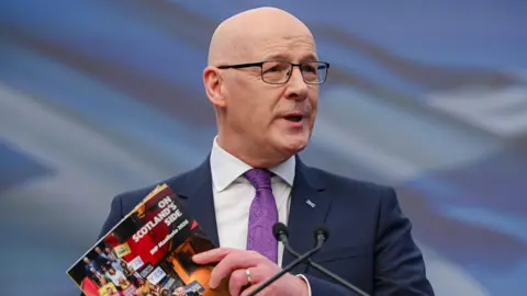 John Swinney holding the SNP's manifesto as he gives a speech in Glasgow 
