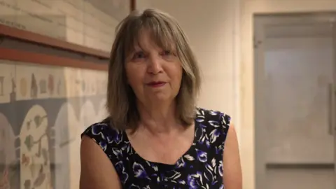 Liz Maiklejohn wearing a black, purple and white patterned t-shirt. She had shoulder length brown hair and is wearing a dark pink lipstick. She is standing in the Bristol Museum in front of the framed tapestry.