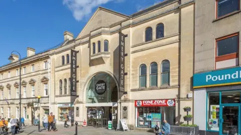 HNW Architects A building with yellow brick with black signs which says 'Emery Gate'. Pedestrians are walking along the pavement in front of it