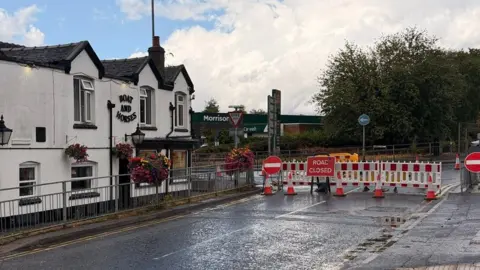 A road with a pub on the corner, across it is a road closed sign and barriers
