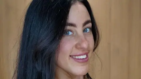 Tia, a young woman, is smiling with her teeth at the camera. She has bright blue eyes and black hair in a side parting and looks at the camera from the side. 