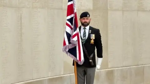 Anthony Frith Anthony Frith standing with the union jack flag. He is wearing a beret and military uniform and white gloves. Behind him is the stone memorial with names on it. 
