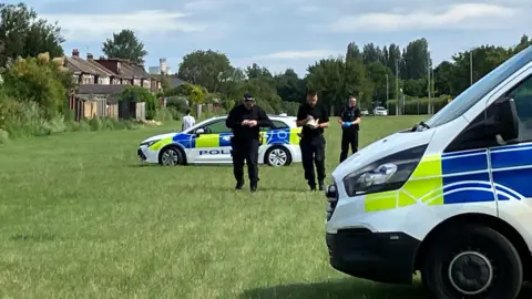Three police officers dressed in black are walking towards the camera. A police van is parked in the foreground with a police car in the background.