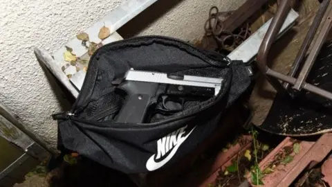 A handgun lying in an unzipped Nike bag, stuffed behind some old furniture in an alleyway