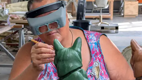 Ama Menec works on the green wax cast of Susie the cat. She is wearing grey goggles and is wearing a sleeveless pink and blue tie-dye dress. To her left is the terracotta-coloured plastolin sculpture.