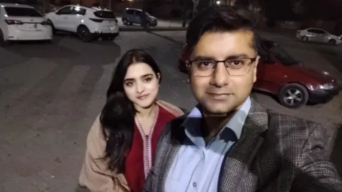 Haleema pictured standing slightly behind Sheharyar in a selfie he is taking of the two in a car park in the dark. She has long black hair and wears a red tunic and beige coat. He wears glasses, a grey checked blazer and a blue shirt