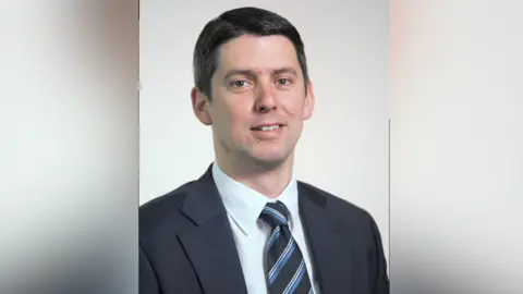 Suffolk County Council A portrait photo of Chris Chambers. He smiles at the camera. He has short black hair and wears a navy suit with a white shirt and black, blue and white tie. 