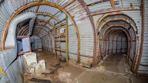 An underground tunnel junction is made of brick and corrugated metal, with arched ceilings and a central pillar, with a second tunnel branching off at a junction in the network of corridors.
