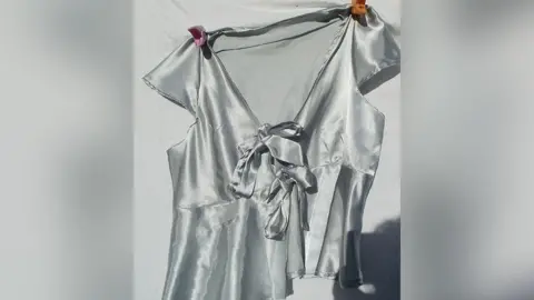 Victoria Ford A silver satin top with bows on the front with a pink peg on the left shoulder and an orange one on the right shoulder. The background is white.