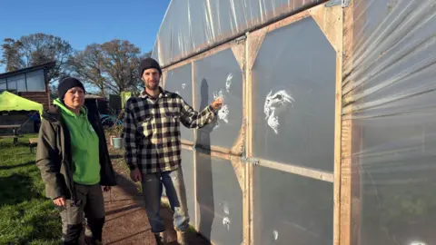 A woman wearing a black woolly hat, green hoodie and a black coat and trousers stands with a man wearing a black woolly hat, a black and white checked shirt and blue jeans. The man rests his hand on the ripped plastic tunnel to his left. 