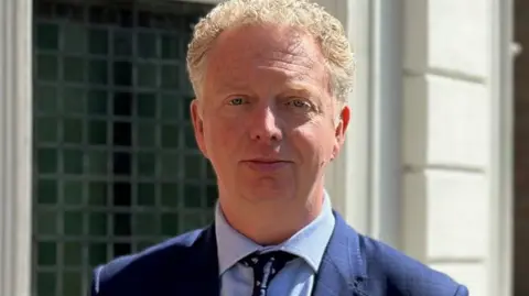 Allinson, who has short fair hair and is wearing a blue suit and tie.