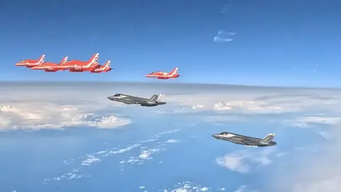 Royal Air Force Five Red Arrows planes fly ahead of two F-35B aircraft, which are painted grey. You can see clouds and the sea below the aircraft. 