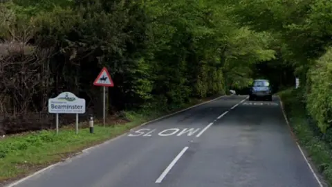 Google street view image of the A3066 - a two-way road lined by thick hedgerows and trees. On the verge is a sign saying Welcome to Beaminster