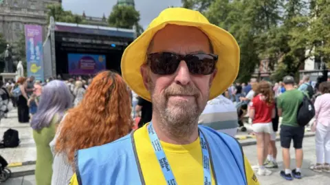 Kevin McVeigh, a man with a yellow hat, dark glasses, a beard, yellow t-shirt and also wearing a blue bib.