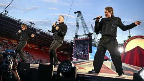 Getty Images The three members of Take That Mark Owen, Gary Barlow and Howard Donald. They are wearing all black and singing on a stage in the centre of the football ground.