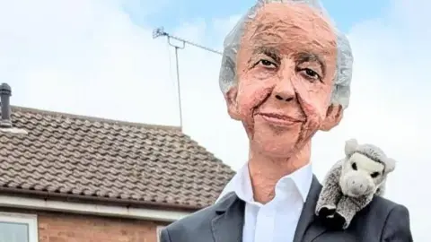 A sculpture depicting Sir David Attenborough wearing a black suit and a white shirt with an animal toy perched on his shoulder. He is perched on a fence which is out of shot. In the background there are some houses.