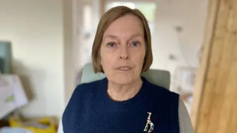 Martin Giles/BBC Woman with bobbed hair, wearing a blue and grey jumper. She is standing in a kitchen.